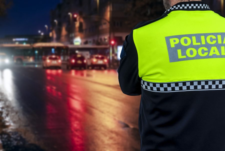 Policía Local de espaldas con chaleco reflectante patrullando de noche en una calle iluminada por los faros de los coches.