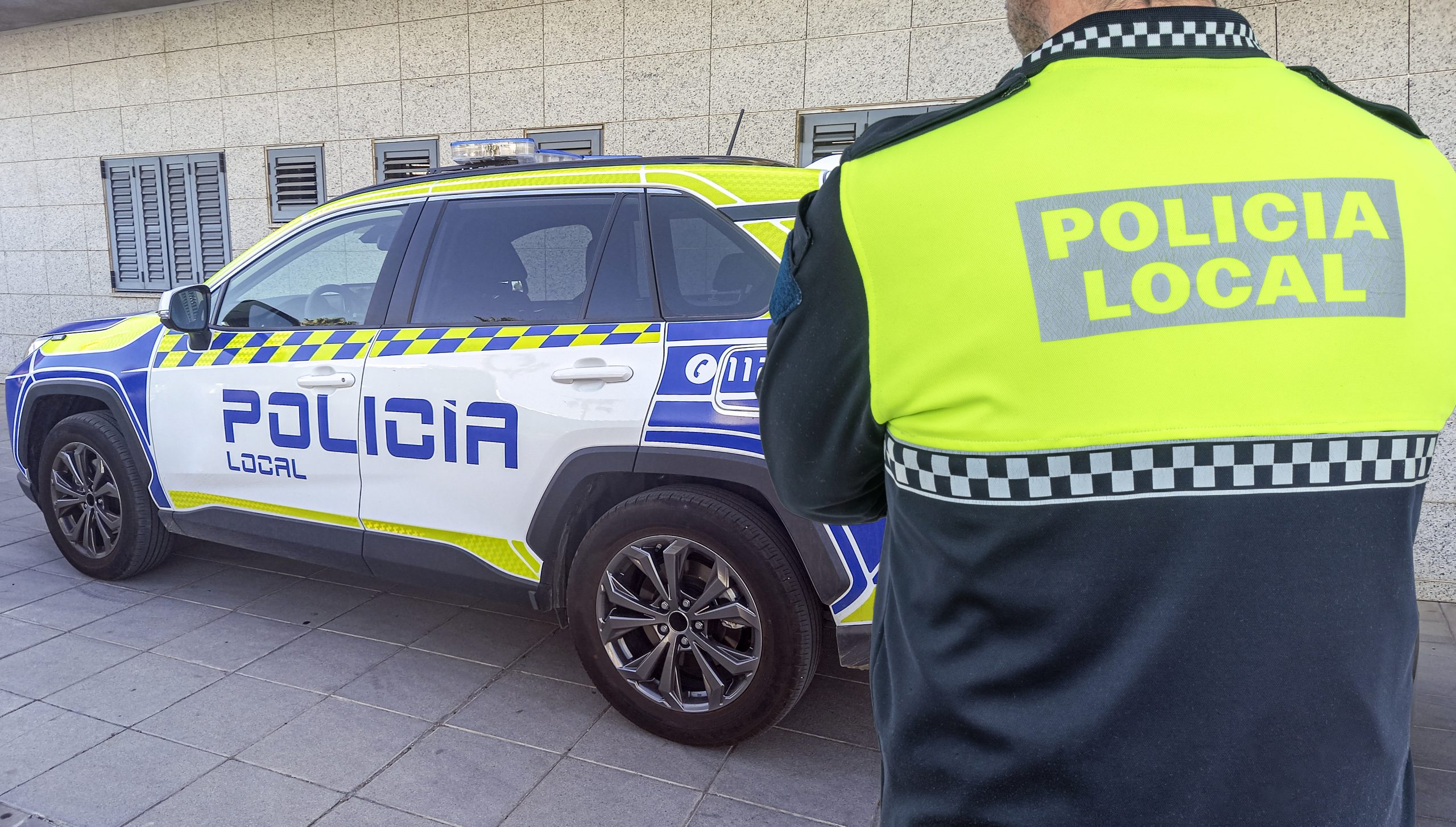 Policía Local de espaldas con chaleco reflectante patrullando de noche en una calle iluminada por los faros de los coches.